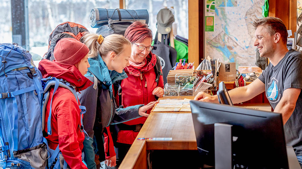 Three hikers with large backpacks have arrived at the service desk of Kilpisjärvi visitor centre. Kilpisjärvi Visitor Centre provides information for tourists, hikers and fishers. Photo: Rami Valonen.